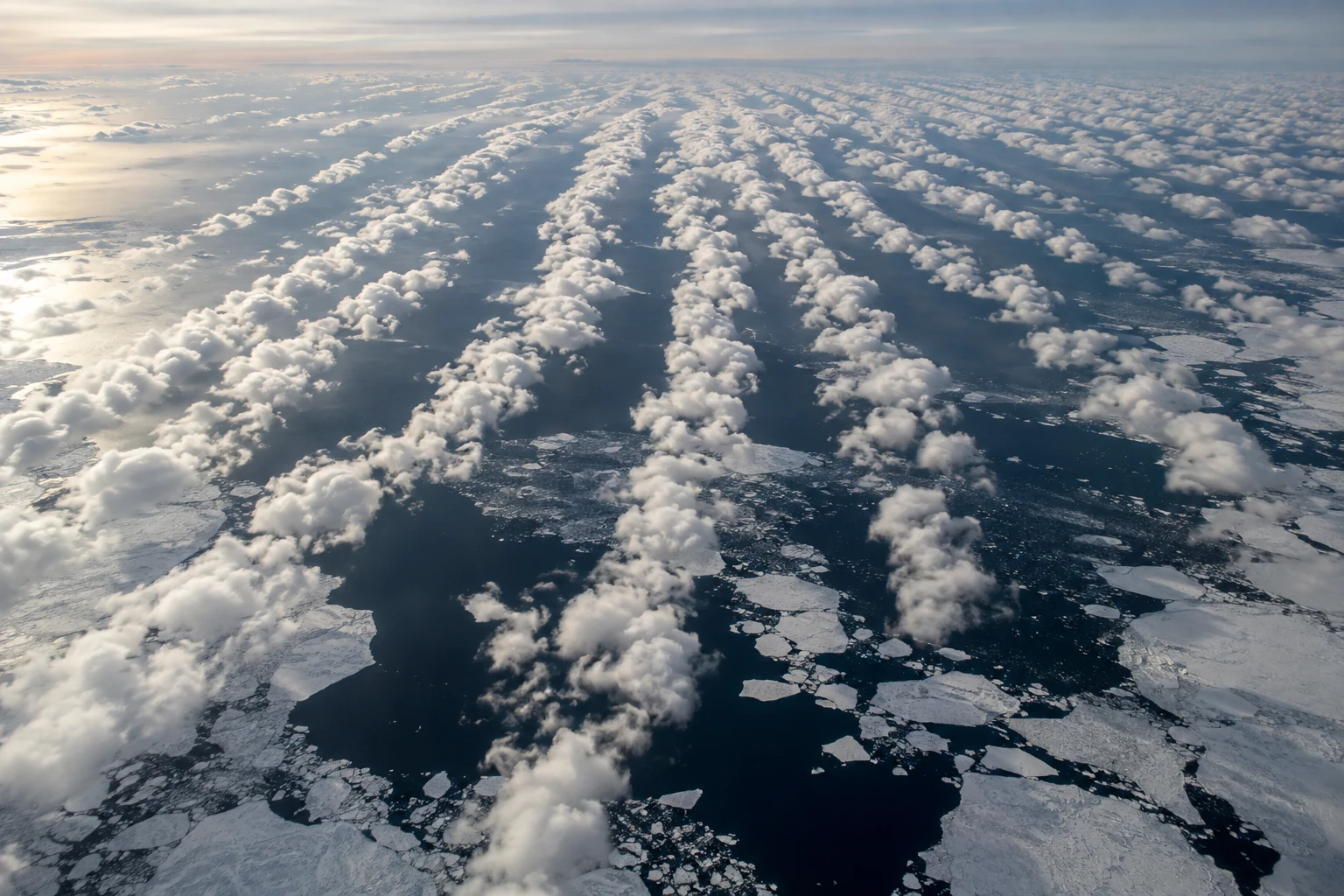 Long parallel rows of white cumulus clouds cross dark Arctic water and broken sea ice under pale winter light.