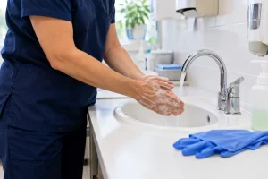 An adult clinician washes bare hands with soap and water beside unused disposable gloves.