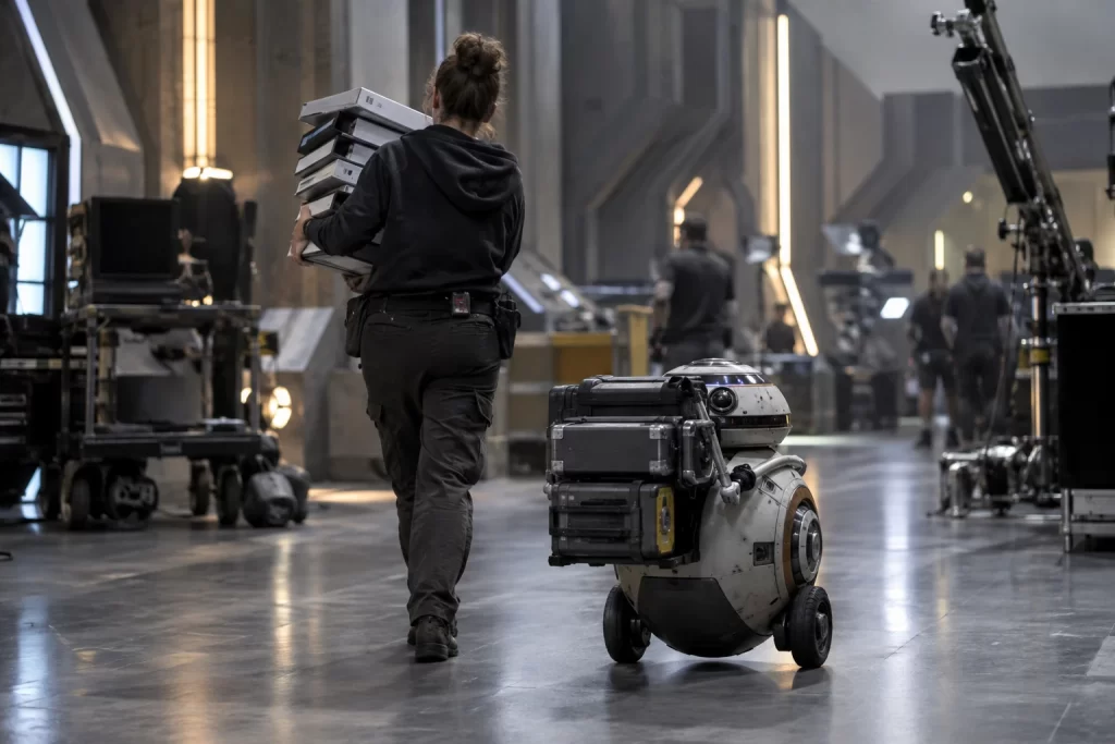 An original service robot follows a production assistant carrying unmarked script binders across a science fiction film set.