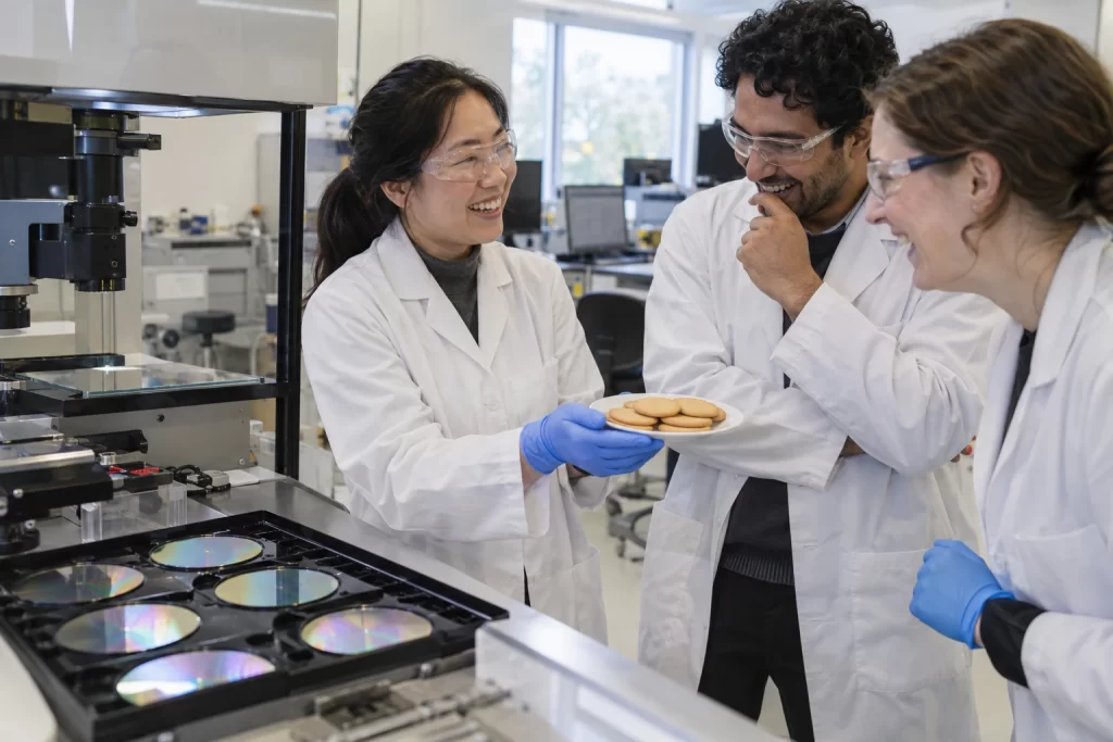 Laughing semiconductor researchers sharing plain biscuits beside a wafer inspection bench