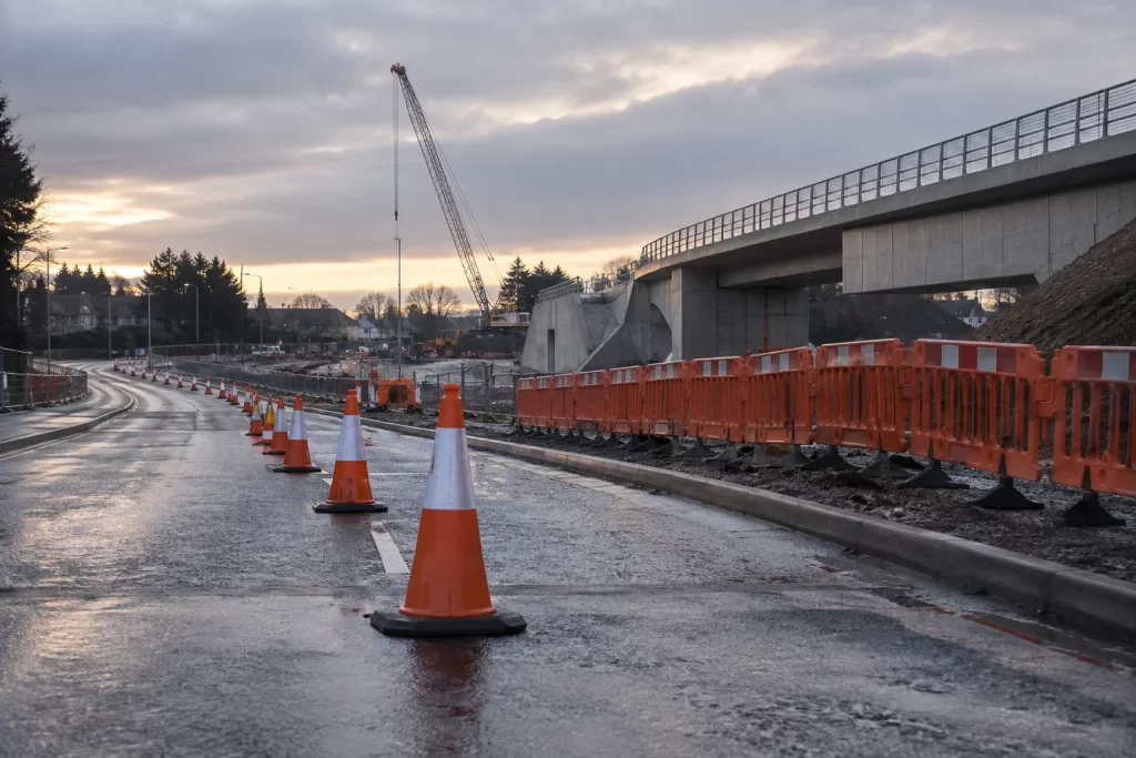 A long line of orange traffic cones and temporary barriers leads past wet roadworks and a new rail bridge at dawn.
